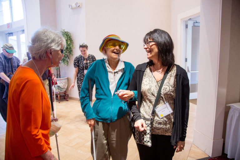 Two women standing. The one one the left is a senior citizen with a white cane