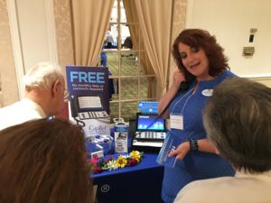 A woman on the right is demonstrating assistive technology to two seniors