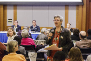 Woman with short grey hair standing in crowd in conference room holding microphone