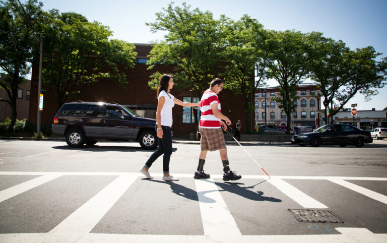 Man with strip shirt and white cane walking in cross walk with O & M specialist trailing