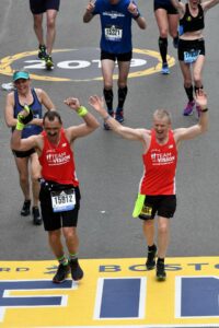 Two men with team with a vision jerseys crossing finish line of boston marathon . Man on left swears bib that says Guide man on right has arms extended up with victory