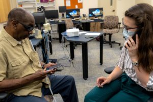 man with sunglasses and white canes by his side, learning how to access features on his phone with an access tech staff member