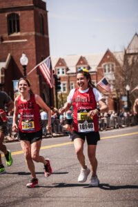 Two runners in red Team With A Vision singlets smiling while running the Boston Marathon together