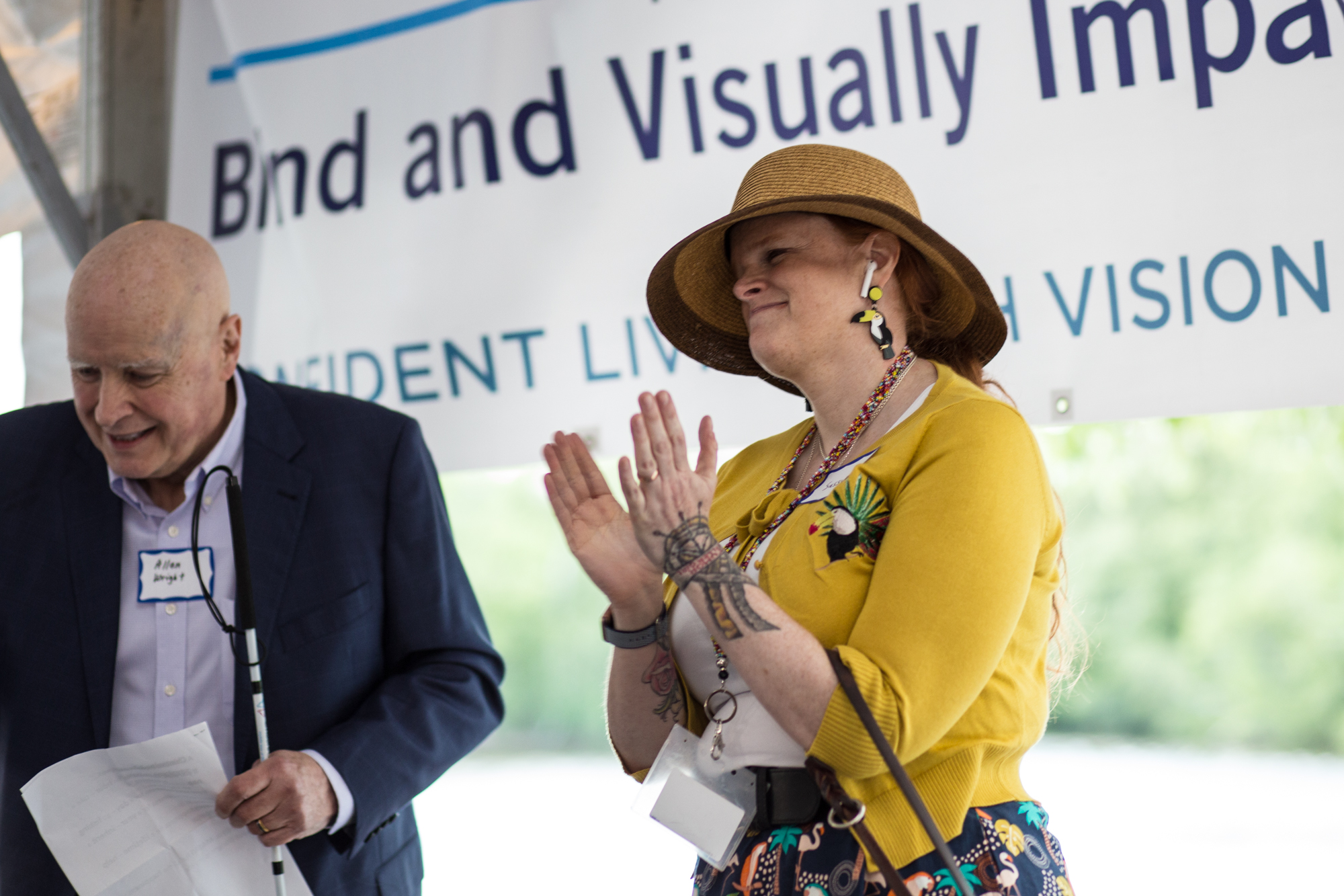 Allen_Wright_and_Sassy Sassy Outwater Wright in a yellow sweater and sunhat clapping beside a Allen Wright in a suit holding papers. A banner behind them reads "Blind and Visually Impaired."