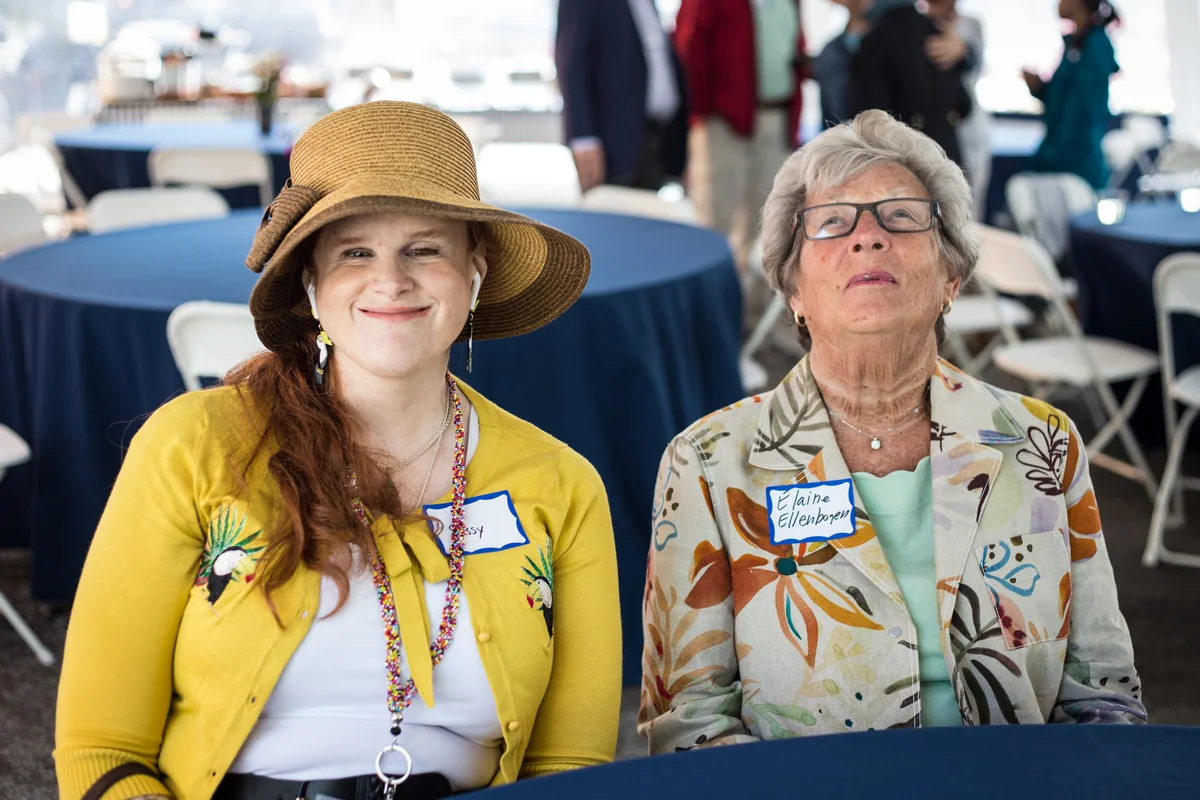 Sassy_and_Elaine Sassy Outwater Wright and Elaine Ellenbogen seated at an event, smiling. Sassy, on the left wears a wide-brimmed hat and yellow cardigan. Ellen, on the right wears a floral blazer.